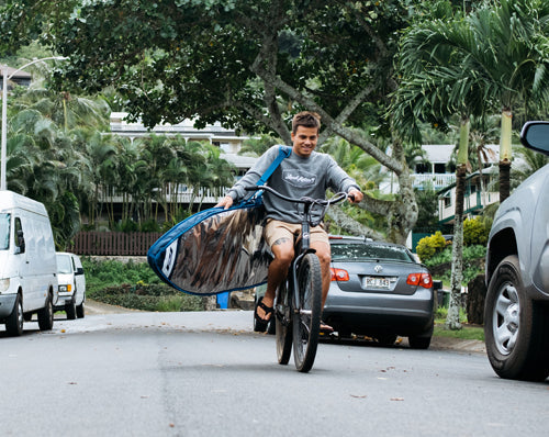 Keanu Asing rides bike with wide ride surfboard day bag.