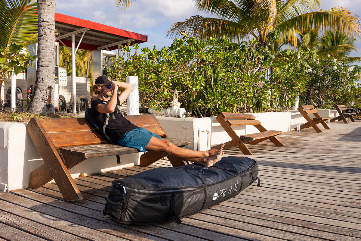 Surfer relaxing on boat dock with feet up on wheeled coffin surfboard travel bag.